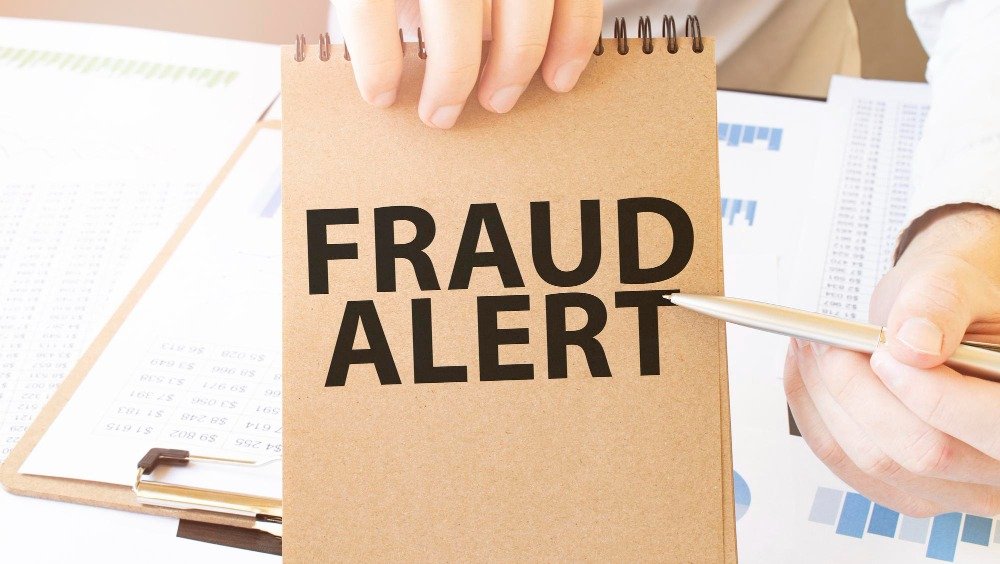 Person holding a notebook with "FRAUD ALERT" written on it, with financial documents in the background.