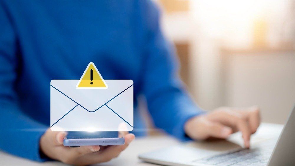 A person in a blue sweater using a smartphone displaying a warning icon over an email envelope, with a laptop in the background.